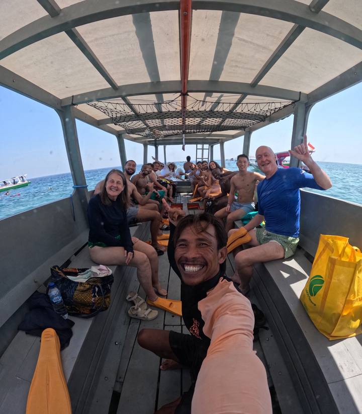 Large snorkelling group smiles and poses on an open boat over clear blue Balinese sea.