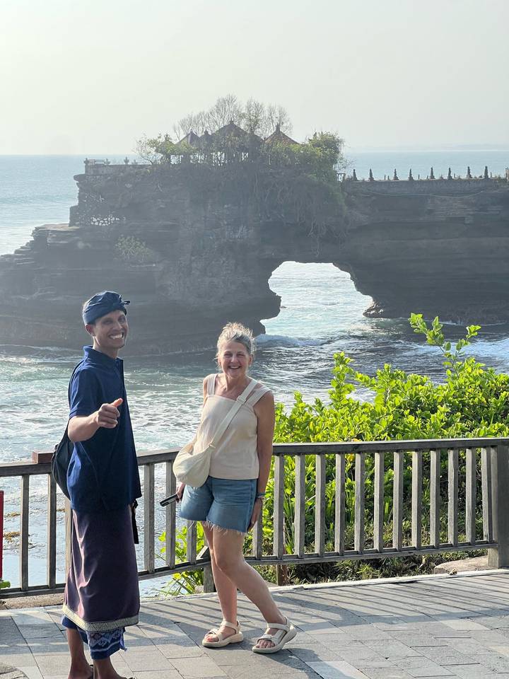 Two travellers smiling in front of an ocean rock arch and temple cliffs with lush greenery.