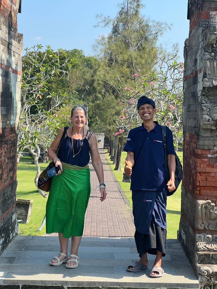Two smiling travellers framed by an ornate Balinese split gate and flowering trees.