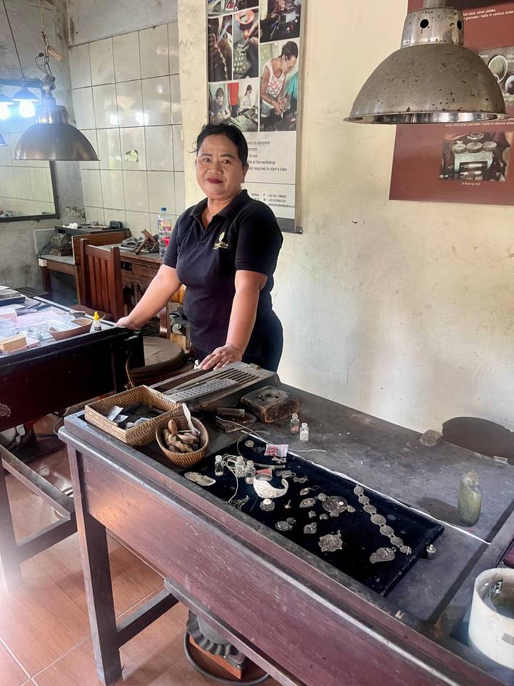 Craftswoman behind a jewellery workbench displaying tools and silver pieces.