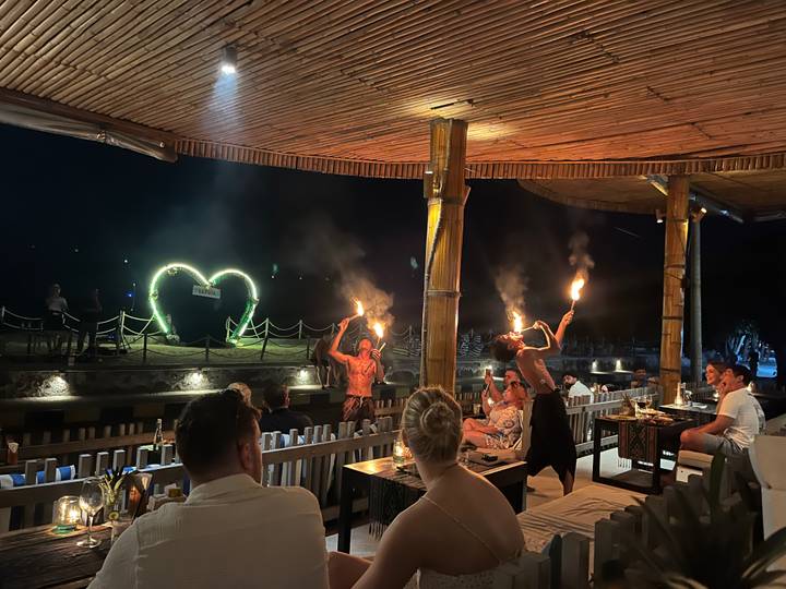 Nighttime fire dancers perform before a heart-shaped light frame at a beachfront restaurant.