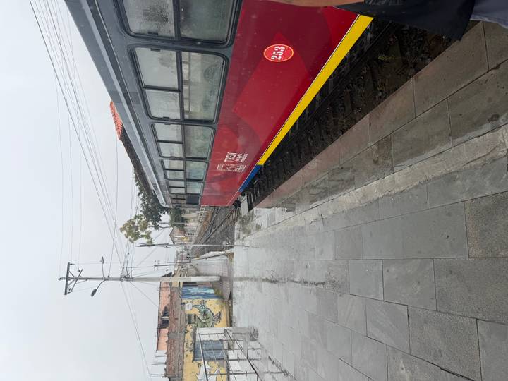 A red and navy Tren Ecuador passenger car sits on wet tracks at a quiet rainy platform.