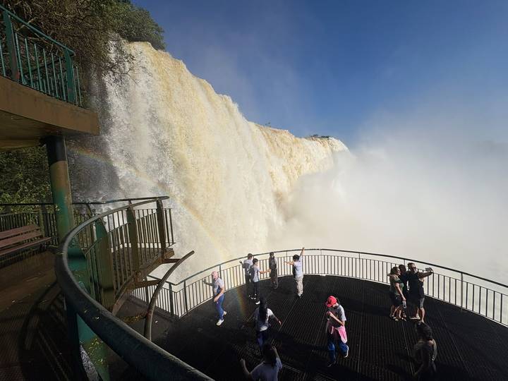 Visitors stand on a spray-drenched balcony beside a towering curtain of Iguazu water with rainbow.