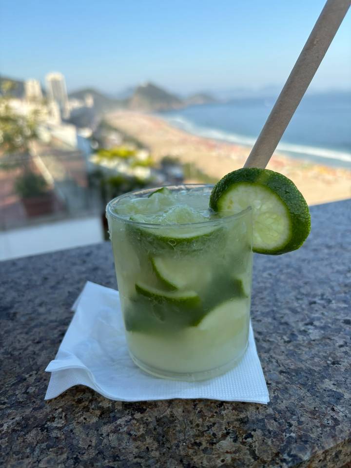 Close-up of a chilled caipirinha cocktail garnished with lime against a blurred beach backdrop.