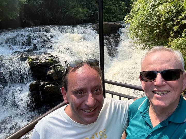 Two men take a selfie in front of a small cascading waterfall surrounded by greenery.
