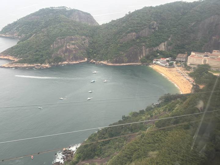 Aerial view through cable-car window of a small beach cove and boats below rocky cliffs.
