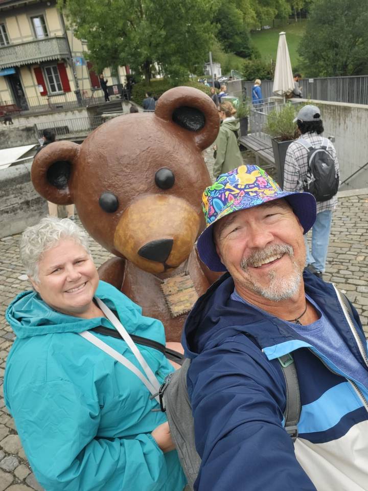 Cheerful couple taking a selfie with a large teddy bear statue in a cobblestone square.