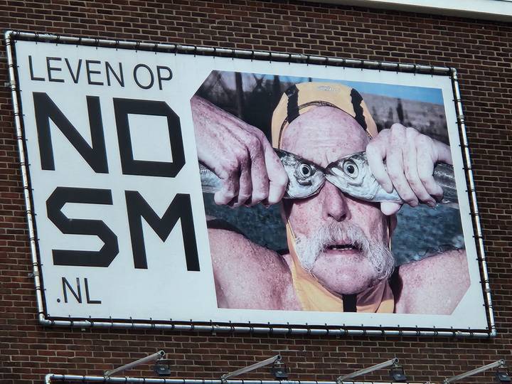 Large outdoor billboard with a humorous photo of a swimmer holding two fish over his eyes and Dutch text.