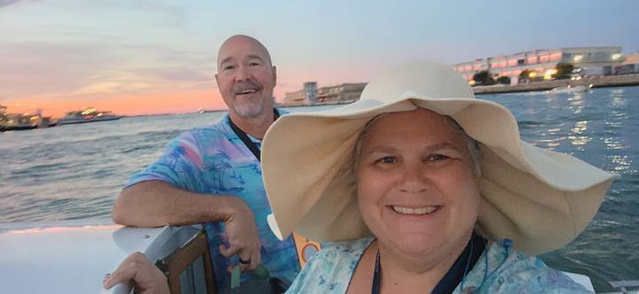 Smiling couple on a boat during a pastel sunset over coastal buildings and calm water.