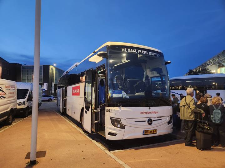 Evening scene of a white tour coach with passengers boarding, lit under blue twilight.