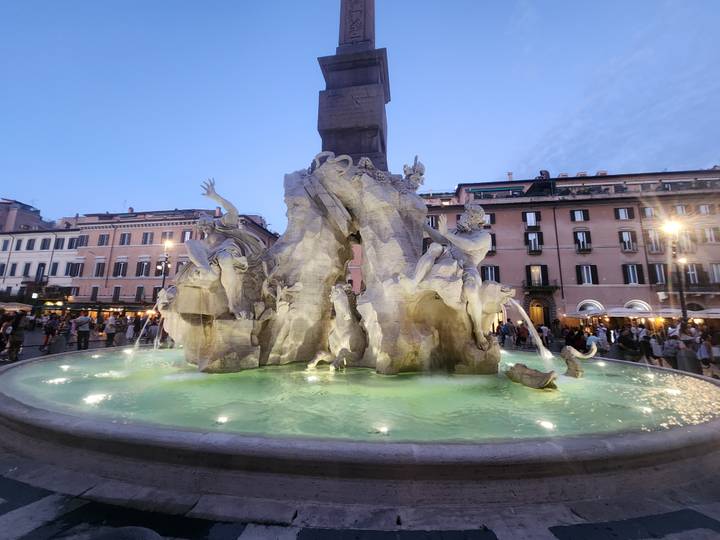 Illuminated Fountain of the Four Rivers in Piazza Navona with surrounding pastel buildings at dusk.