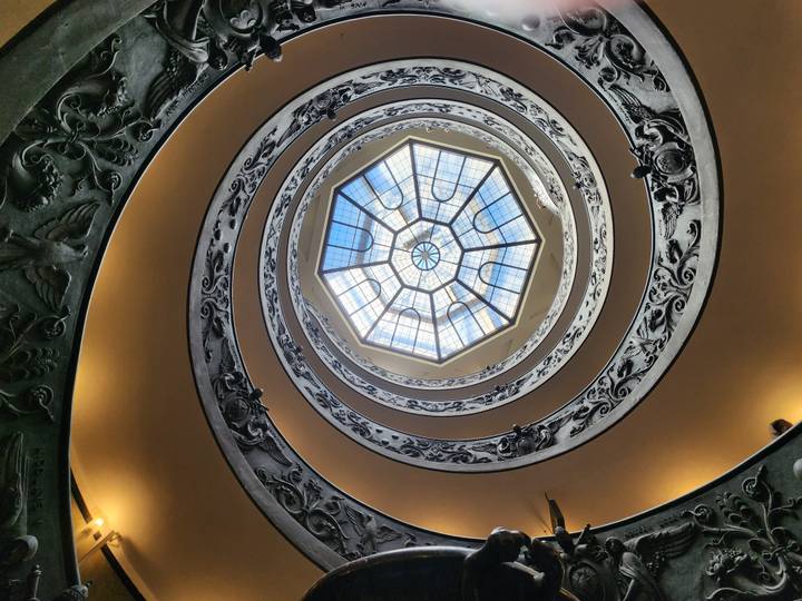 Looking up a spiraling ornate staircase with a stained-glass skylight at the Vatican Museums.