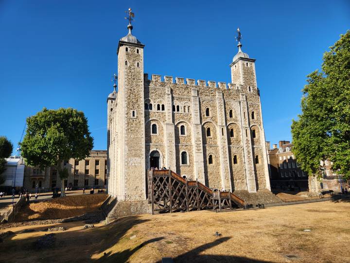 The White Tower of London rises against a clear cobalt sky beside lush green trees.