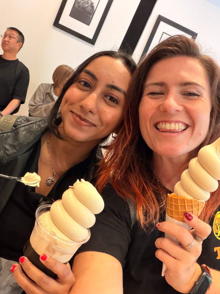 Two friends smile for a close-up selfie while enjoying tall swirled ice-cream cones indoors.