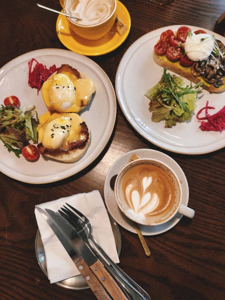 Flat-lay of brunch plates with eggs Benedict, salad and a latte with heart-shaped foam art.