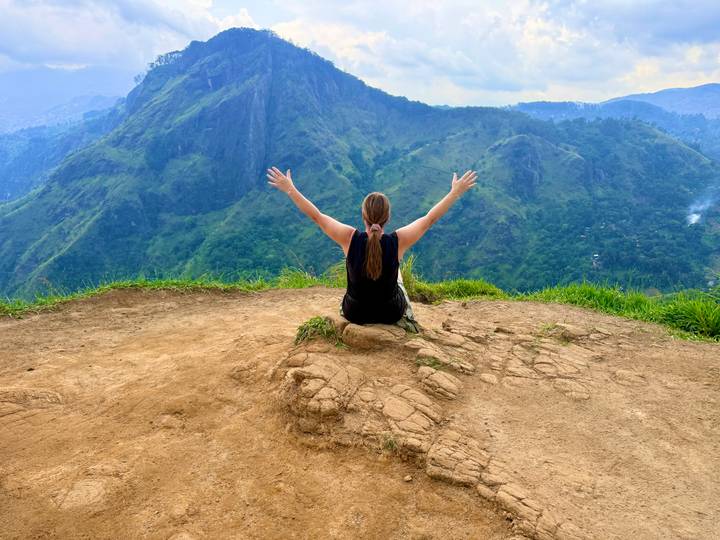 Traveller sitting with arms raised in triumph on a cliff top overlooking verdant mountains.