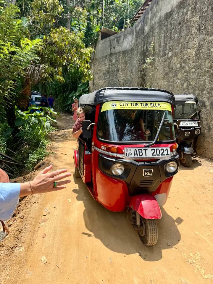 Bright red tuk-tuk on a dusty lane with passengers and Sri Lankan licence plate.