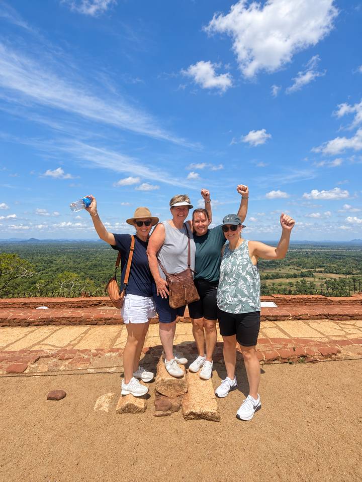 Four friends on a stone terrace raising their arms in celebration over a vast green landscape.