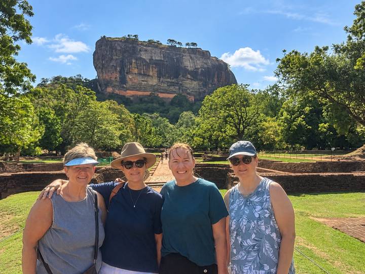 Four women posing in front of the massive Sigiriya Rock surrounded by lush greenery under a bright blue sky.