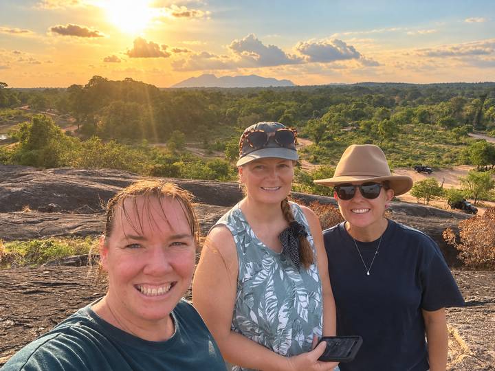 Three women smiling on a rocky viewpoint at golden sunset with forested plains and distant hills behind them.