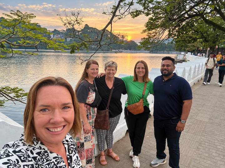 Five travelers standing by a tranquil lakeside promenade at sunset with town lights beginning to glow.