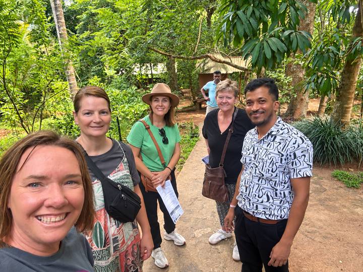Travel group with their guide smiling on a shaded garden path surrounded by tropical plants.