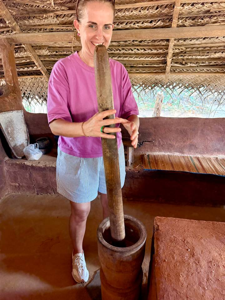 Person in casual clothes using a long wooden pestle inside a rustic mud-brick hut with thatched roof.