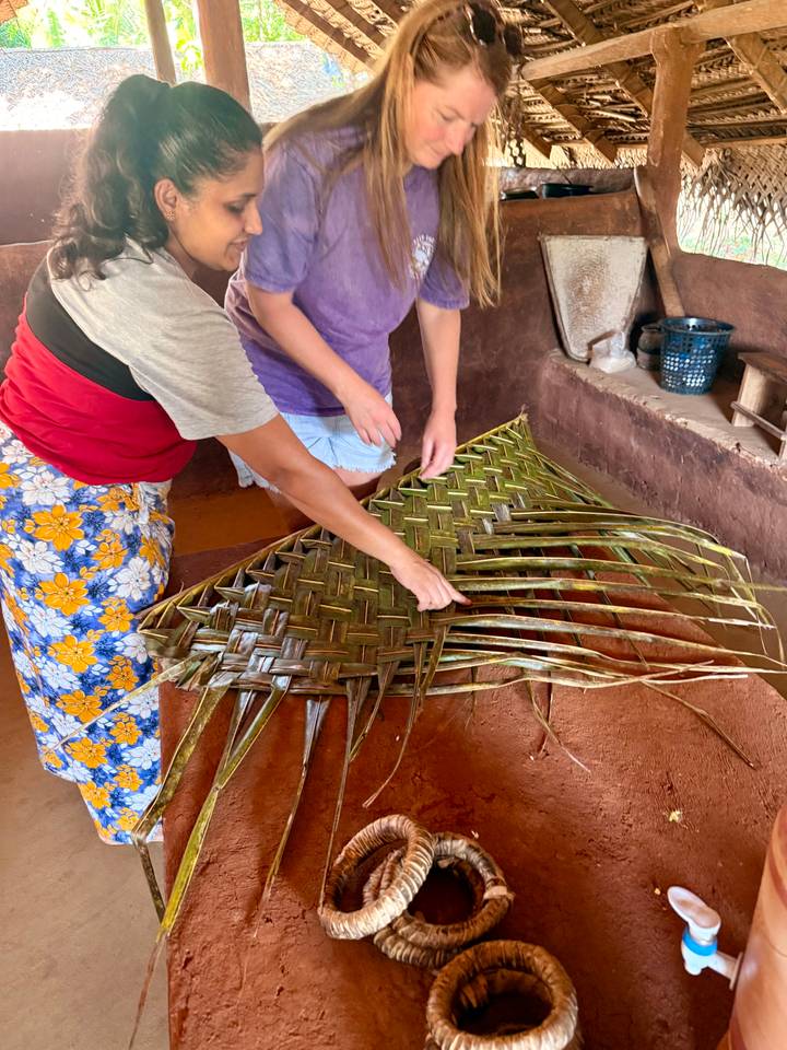 Two people weaving palm fronds into a patterned mat on an earthen floor workshop.