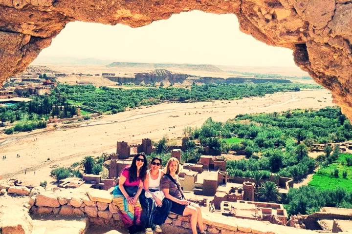 Three travelers sit beneath a natural stone arch overlooking the fortified village and lush valley of Ait Benhaddou.