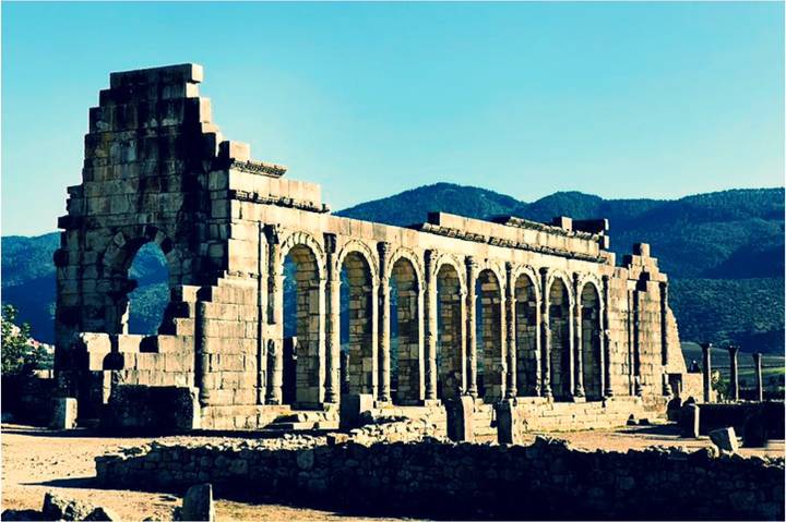Ruined Roman arches of Volubilis standing against a backdrop of green mountains under clear sky.