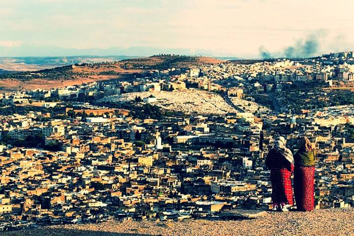 Two women in traditional dress overlooking the sprawling old cityscape of Fes from a high viewpoint.