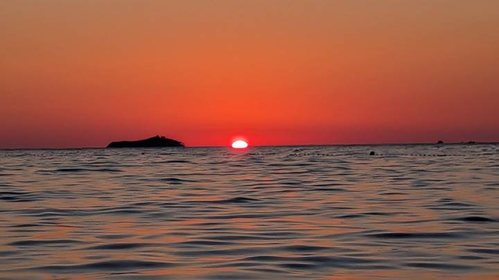 Fiery red sun setting into calm Adriatic Sea with small island silhouette on horizon.