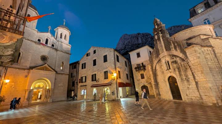 Evening scene of stone church and old buildings in Kotor’s old town square with soft blue twilight sky.