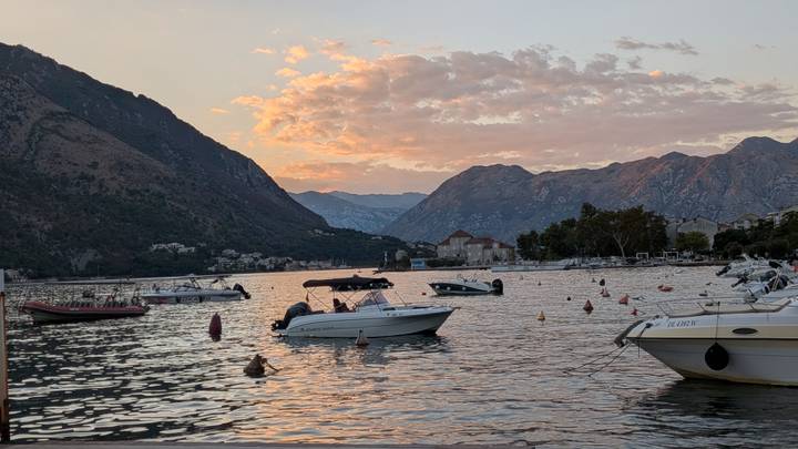 Boats anchored on tranquil Bay of Kotor with pastel sunset clouds over surrounding mountains.