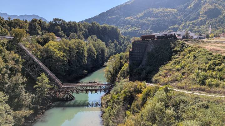Old wooden railway bridge spans a green river gorge amid thick forested mountains.