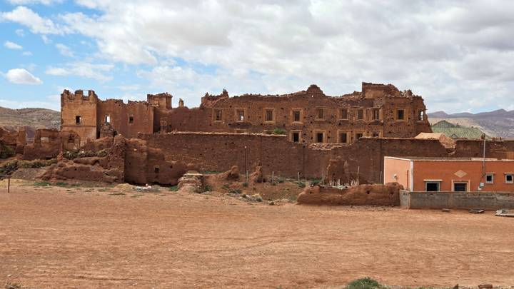 Ruined earthen kasbah walls and empty windows stand in a barren landscape under a partly cloudy sky.