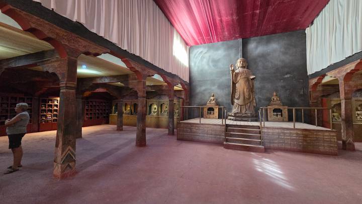 Dimly lit interior hall exhibits statues and paintings with a large Buddha figure on a platform.