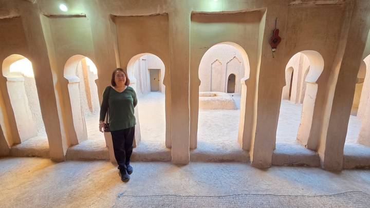 Woman stands inside an ancient adobe kasbah corridor with repeating arches and soft golden light.