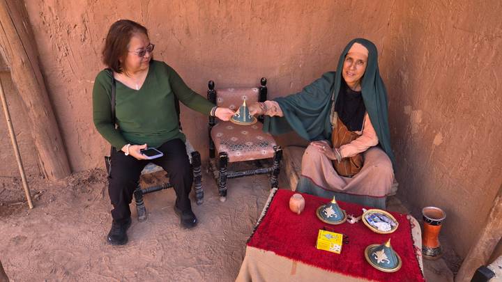 Local host in green veil offers tea to a tourist seated beside her against an earthen wall.