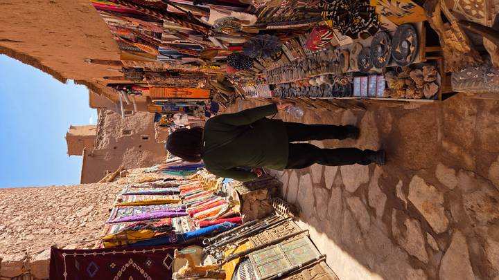 Narrow souk alley lined with colorful rugs, jewelry and souvenirs, tourist walking away through sunlight.