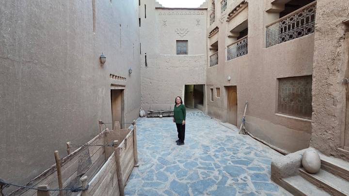 Woman stands in a narrow stone-paved kasbah alley flanked by high adobe walls and wooden balconies.