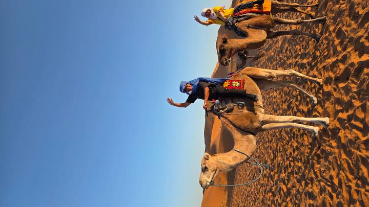 Two travelers in bright clothing wave while riding camels across golden Sahara dunes under a clear sky.