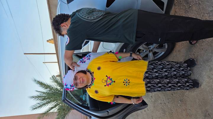 Local guide helps tourist adjust traditional attire beside a parked vehicle in desert village.