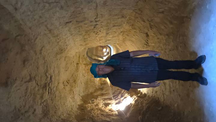 Man wearing blue scarf stands in a narrow underground tunnel carved from earth, light filtering in.
