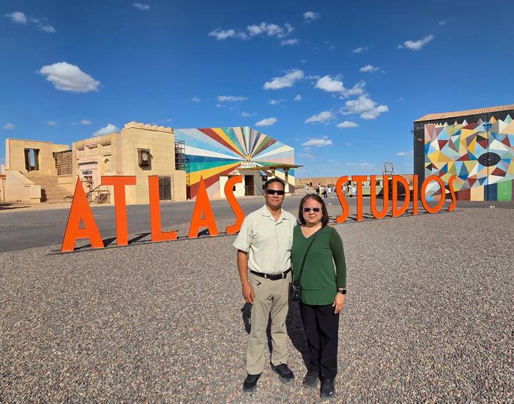 Couple stands in front of large orange ATLAS STUDIOS sign and colorful film sets under blue sky.