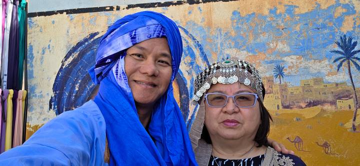 Two travellers in traditional headwear smiling in front of a colorful desert-themed mural.