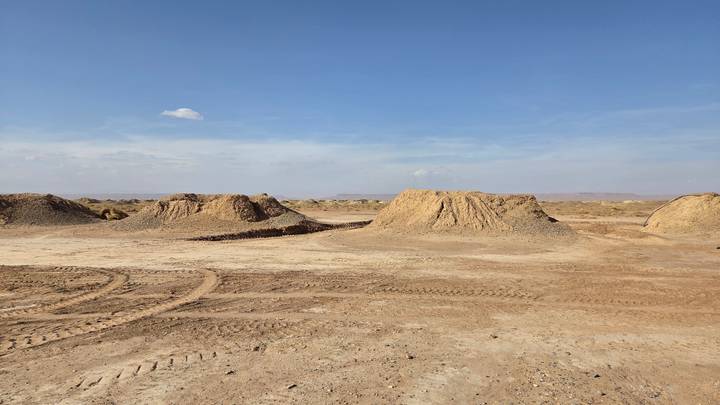 Barren desert landscape with low earthen mounds and tyre tracks under clear sky.