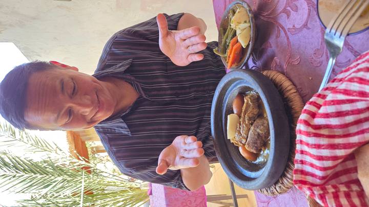 Smiling diner presenting a steaming tajine dish at an outdoor restaurant table.