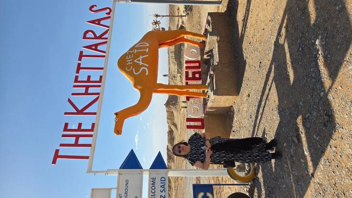 Visitor standing beside bright camel statue under sign 'The Khettaras' in desert area.