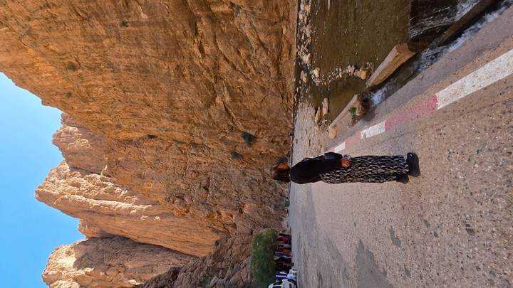 Visitor gazing up at towering orange cliffs beside shallow stream in a canyon road.